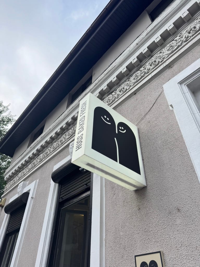A projecting square sign on a gray stucco building reads “HUGS BAKED FRESH” along the edge and shows a minimalist black graphic of two rounded shapes with smiling faces, resembling a hug. Decorative white moldings frame the windows and cornice; trees and cloudy sky are visible above.