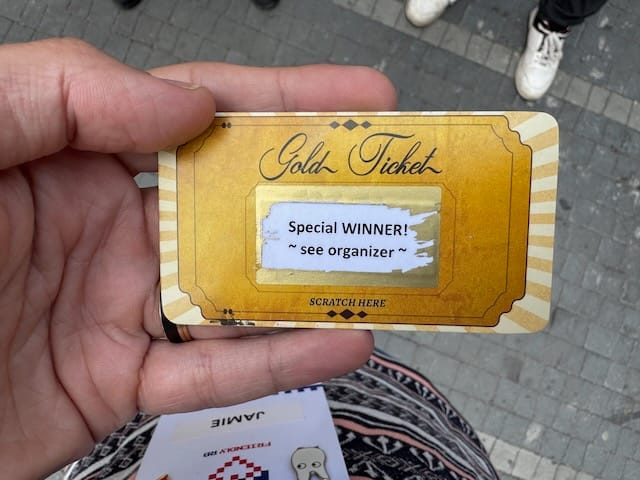 A hand holds a golden raffle-style card labeled “Gold Ticket,” with a scratched panel revealing the message “Special WINNER! ~ see organizer ~.” The card edges have vintage detailing, and a lanyard badge and feet on paving stones are visible in the background.