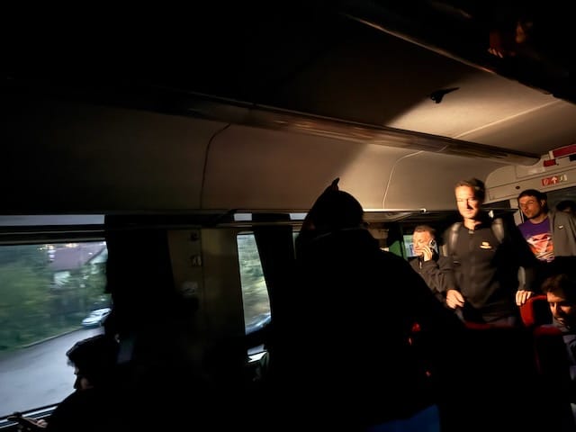 Dim, unlit train car where a beam of sunlight and a bright phone torch cut through the darkness. Passengers file into the aisle with bags, faces partly lit by the torch while the rest of the seats and ceiling remain in shadow.