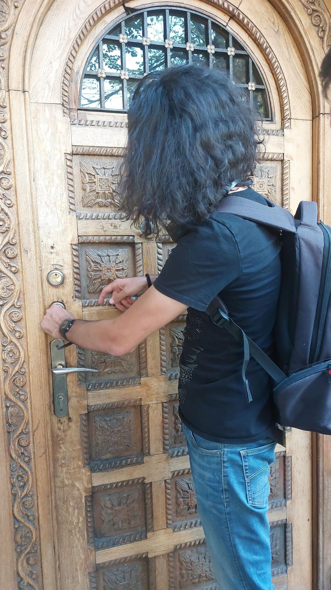 A person with long wavy hair, a black T‑shirt, jeans, and a black backpack stands at an ornate carved wooden door, focusing on the lock with both hands near the keyhole. The door has detailed square panels and an arched window with black metal grilles at the top.