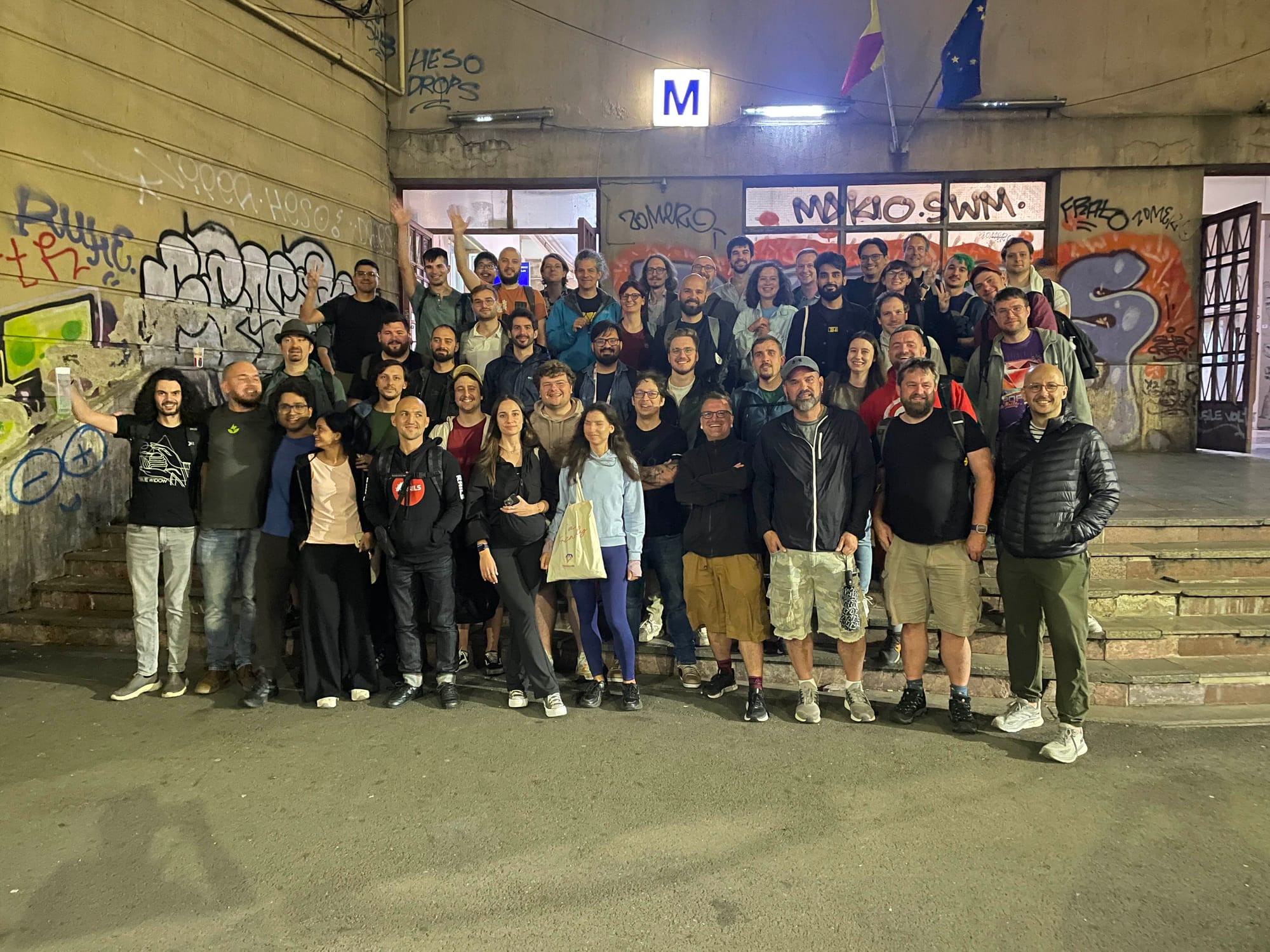 A large group of smiling people poses on the steps outside a graffiti‑covered building at night. An illuminated “M” sign and two flags hang above the entrance. The crowd fills the stairs and doorway, with backpacks and casual jackets, under warm artificial light.