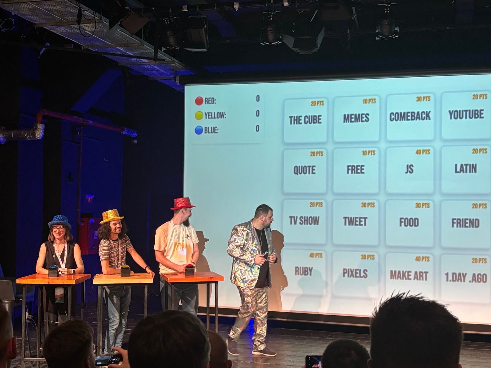 On a stage set up like a game show, three contestants in colorful glittery hats stand at podiums while a host in a shiny patterned suit faces a giant screen. The projected board shows categories with point values, including “THE CUBE,” “MEMES,” “COMEBACK,” “YOUTUBE,” “QUOTE,” “FREE,” “JS,” “LATIN,” “TV SHOW,” “TWEET,” “FOOD,” “FRIEND,” “RUBY,” “PIXELS,” “MAKE ART,” and “1.DAY.AGO.” A scoreboard at left lists Red, Yellow, and Blue teams at zero as the audience watches.