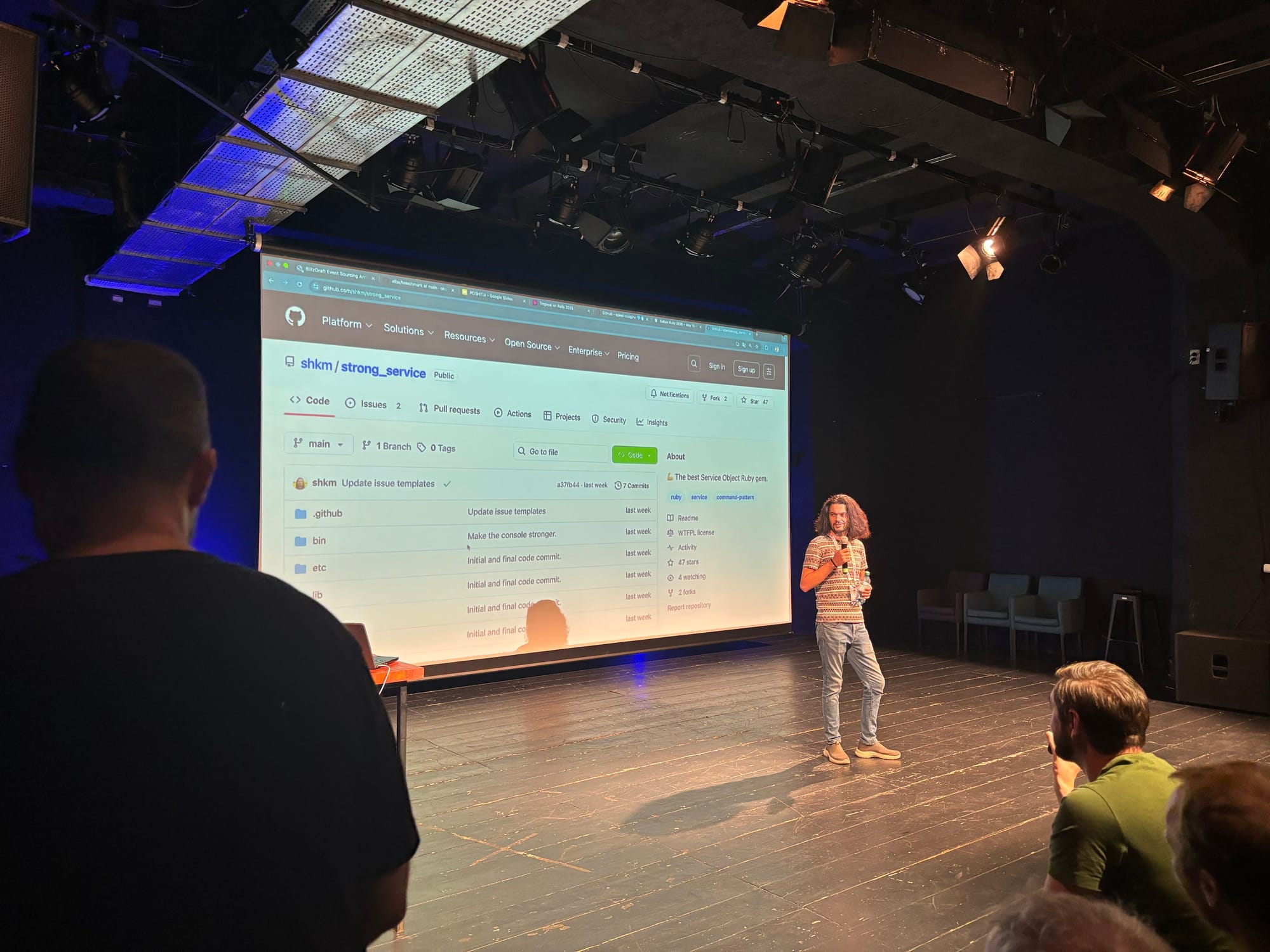 A speaker on a dimly lit stage presents in front of a large projected browser window showing a GitHub repository titled “shkm/strong_service.” The audience sits in the foreground, and the presenter stands near center with a microphone, casual attire, and curly hair. Stage lights and ceiling rigging are visible above; a few chairs line the right side of the stage.