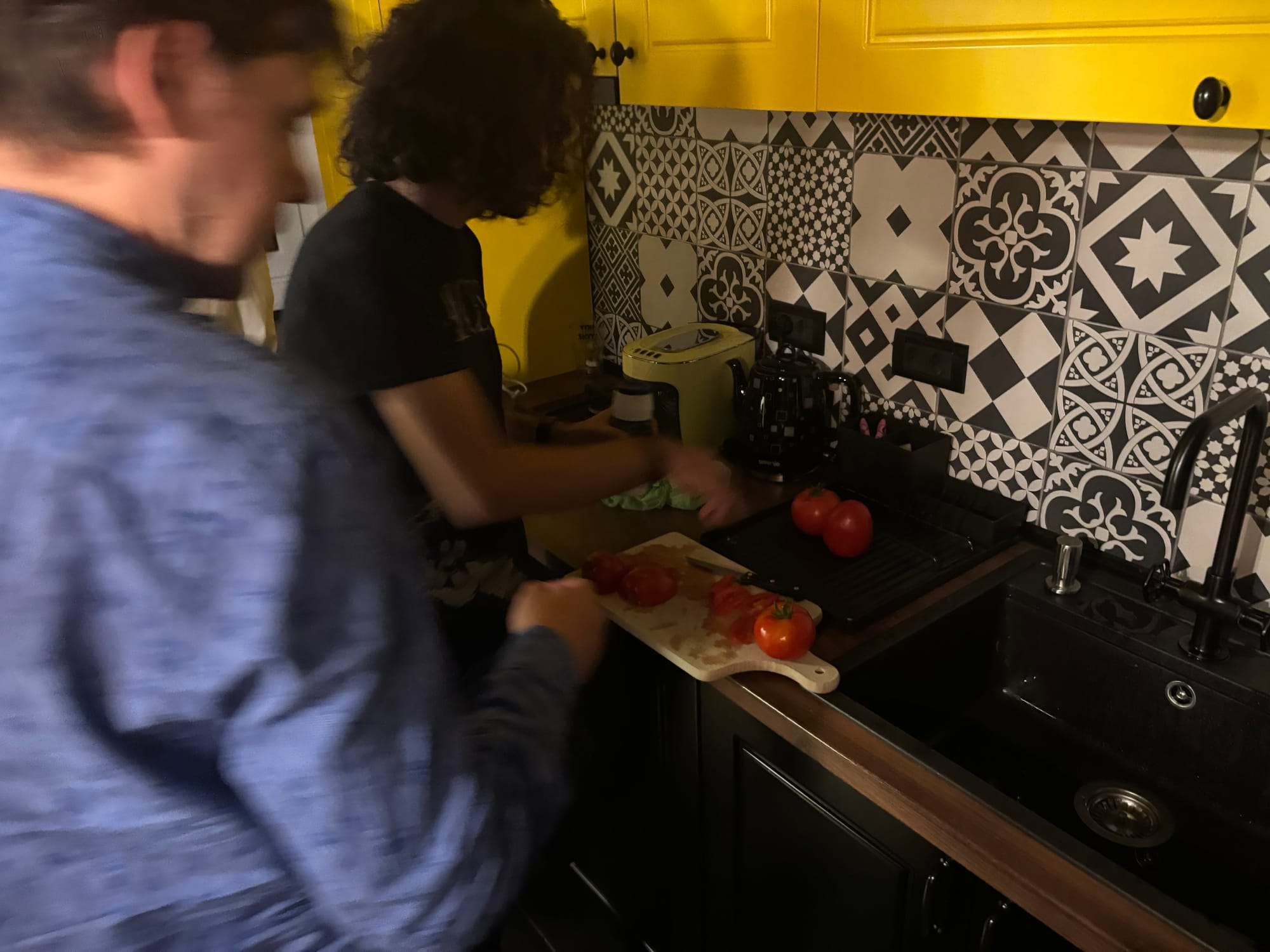 Two people in a cozy kitchen slice and sample tomatoes at the counter. One person in a dark shirt is cutting tomatoes on a white board beside a sink, with whole tomatoes resting nearby. The foreground shows another person in a blue patterned shirt reaching toward the board. The backsplash features black‑and‑white geometric tiles, and bright yellow cabinets run overhead alongside a black faucet and appliances.
